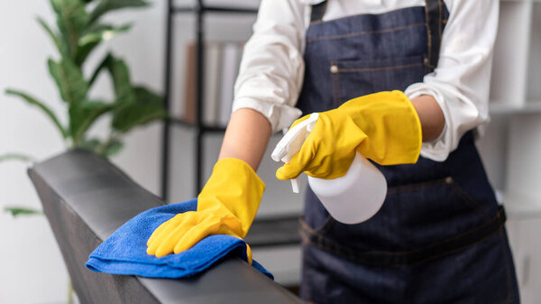 Домохозяйка в фартуке в перчатках для распыления гигиенического спрея на диван и использования микроволоконной ткани для очистки мебели во время работы и уборки в доме.