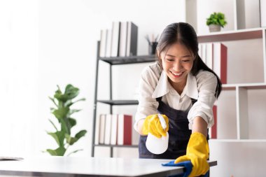 Masaya sprey sıkmak için eldiven giyen önlüklü bir ev hanımı ve evde çalışırken temizlik yaparken temizlik yapmak için mikrofiber kumaş kullanıyor..