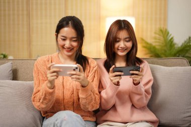 Genç lezbiyen çift rahat koltukta oturuyor ve akıllı telefon kullanarak kız arkadaşlarıyla video oyunu oynuyorlar. Bu arada birlikte oturma odasında vakit geçiriyorlar..