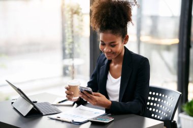 Takım elbiseli Afrikalı Amerikalı iş kadını elinde kahve fincanı tutarken ve akıllı telefondan muhasebe bilgilerini okurken aynı zamanda modern çalışma alanında evrak işleriyle uğraşan bir tablet üzerinde çalışıyor..
