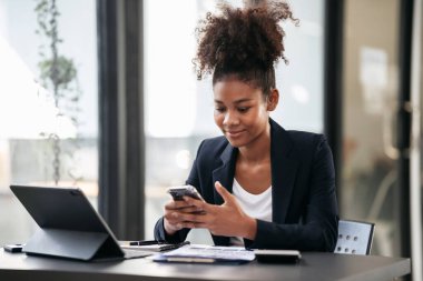 Takım elbiseli Afrikalı Amerikalı iş kadını akıllı telefondan muhasebe bilgilerini okuyor ve modern çalışma alanında evrak işleriyle uğraşırken projenin stratejisini düşünüyor..