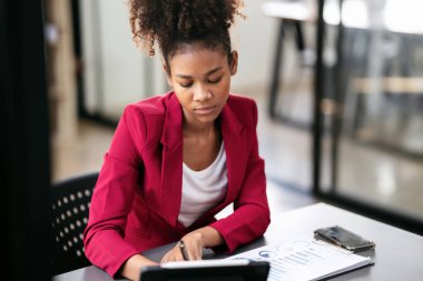Takım elbiseli Afrikalı Amerikalı iş kadını, iş dünyasının muhasebe bilgilerini tablet ve evrak işleriyle ilgili bilgileri okuyordu ve modern çalışma alanında proje stratejisi üzerinde çalışırken ve düşünürken deftere not alıyordu..