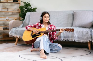 Genç bir kadın gitar çalıyor ve müzik dinlemek için kulaklık takıp şarkı söylüyor ve evdeki oturma odasında yerde oturuyor..
