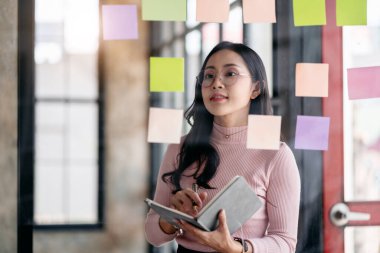 İş planlama ve ortak fikir kavramı, iş kadını cam duvardaki notlarda bilgi okuyor ve ofiste iş planlaması hakkında düşünürken ve analiz yaparken not defterine bilgi yazıyor..