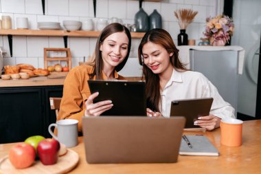 İki genç kadın iş bilgilerini pano ve dijital tablette birlikte okuyarak yeni proje işiyle ilgili beyin fırtınası yaparken aynı zamanda evde mutfaktaki masada dizüstü bilgisayarla çalışıyor..