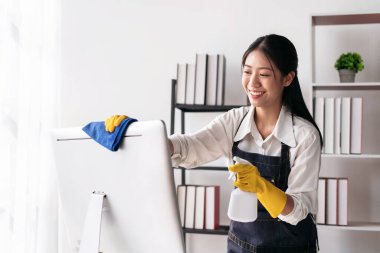 Önlüklü ev kadını, bilgisayara sprey sıkmak için eldiven giyiyor ve evde çalışırken temizlik yaparken temizlik yapmak için mikrofiber kumaş kullanıyor..