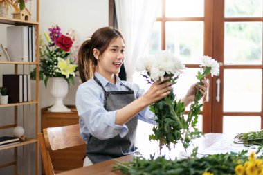 Önlüklü kadın çiçekçi elinde beyaz kasımpatı ile çiçekçi dükkanındaki masanın üzerindeki çiçek buketini düzenlemek için çiçek tasarlıyor..
