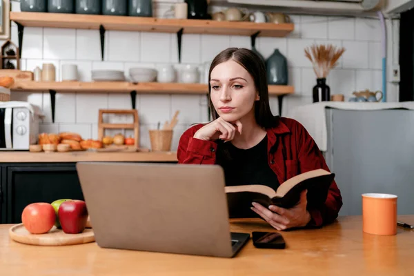 年轻的女商人坐在厨房里 一边看书 一边阅读笔记本电脑上的商业信息 一边思考新商业计划的策略 — 图库照片