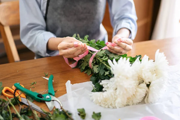 Female florist in apron using pink ribbon to tie for creating and making flower bouquet of white chrysanthemum in her flower shop for delivery to customer.