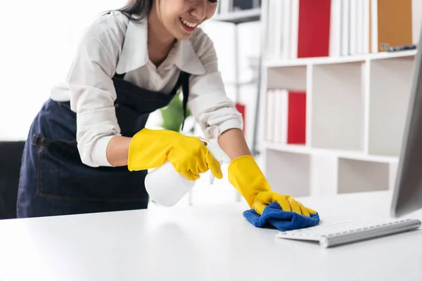 Housewife in apron wearing gloves to spraying hygiene spray on computer and using microfiber fabric to wiping cleanup the table while working and cleaning furniture in the house.
