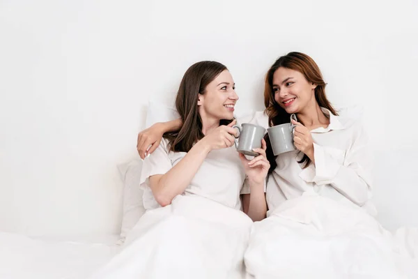 stock image Young asian couple woman clinking cup and drinking chocolate cup while embracing and lying under blanket and leaning on comfortable pillows on the bed in bedroom at home.