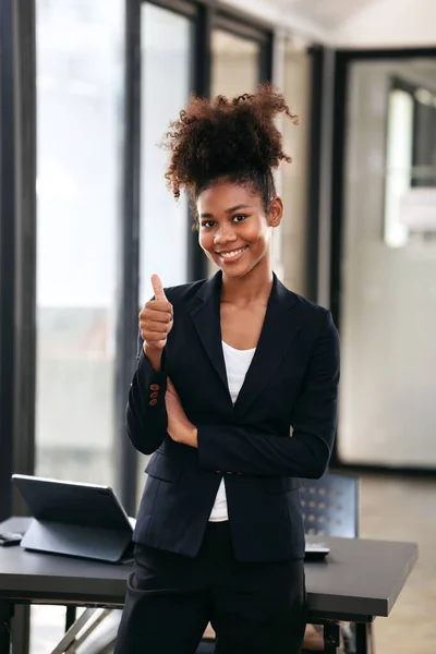 African american businesswoman in suit standing to making thumb up and smiling on camera after successfully of working about accounting of new business on tablet with paperwork in modern workspace.