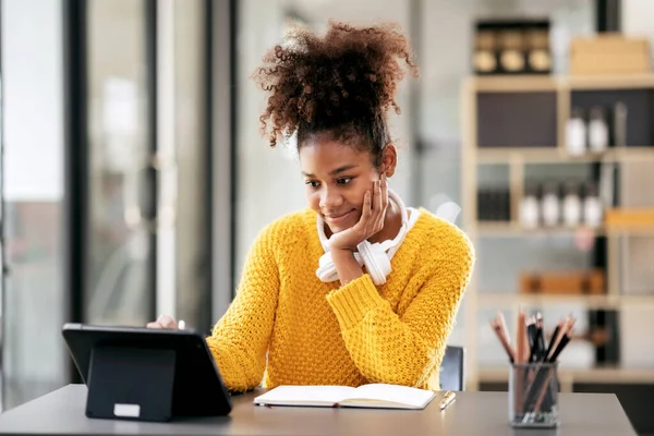 African american student woman in sweater watching e-learning to studying lesson online class on tablet and taking notes while sitting on the desk to learning knowledge and education in university.