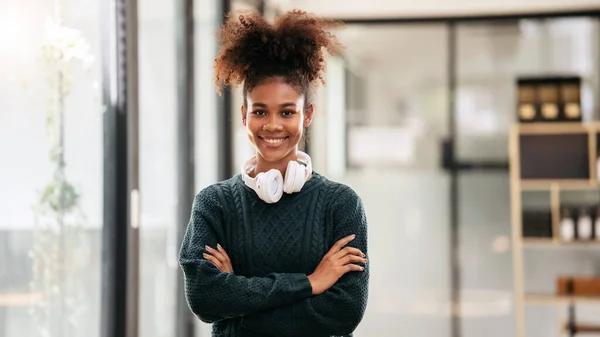 African american student woman in sweater wearing headphones around neck and standing with arms crossed after studying lesson online class while learning education and knowledge in university.