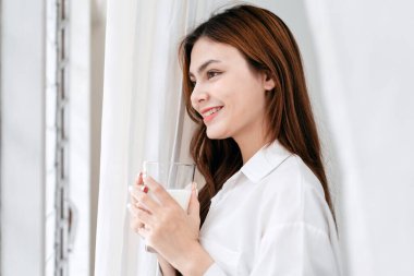 Evde eğlence yaşam tarzı konsepti, günlük giysiler içinde, pencerenin yanında duran, rüya gibi süt içen, evde yaşam tarzıyla eğlenen genç bir kadın..