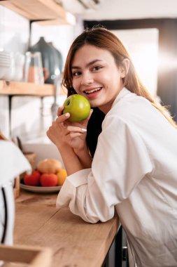 Ev eğlencesi yaşam tarzı konsepti, mutfak odasının yanında günlük giysiler içinde duran ve evde yaşam tarzıyla eğlenirken sağlıklı yemekle birlikte yeşil elma meyvesi yiyen genç kadın..
