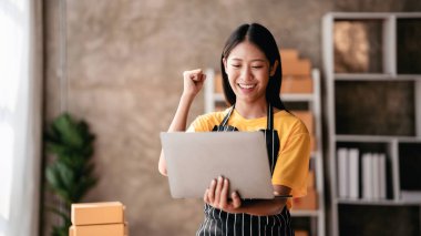 Küçük işletme paket teslimatı konsepti, çevrimiçi siparişleri kontrol eden kadın girişimci ve çevrimiçi satış işlerinde çalışırken başarılı bir şekilde kutlama yapmak için kollarını kaldırma.