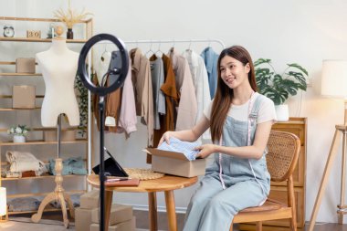 Kadın çevrimiçi satıcı moda tanıtımı için akıllı telefon kullanıyor ve müşteri göndermeye hazırlanmak için karton kutulara ürün paketlerken çevrimiçi kıyafet satıyor..