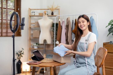 Kadın çevrimiçi satıcı moda tanıtımı için akıllı telefon kullanıyor ve müşteri göndermeye hazırlanmak için karton kutulara ürün paketlerken çevrimiçi kıyafet satıyor..