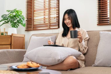 Kadın girişimci büyük rahat koltukta oturuyor ve kahve içerken ve evde oturma odasında çalışırken veri yazmak için yastığa tablet koyuyor..