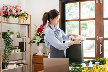 Çiçekçi dükkanında çiçek buketi hazırlarken ve dizayn ederken beyaz kasımpatı kağıt torbada düzenleyen önlüklü kadın bir çiçekçi..