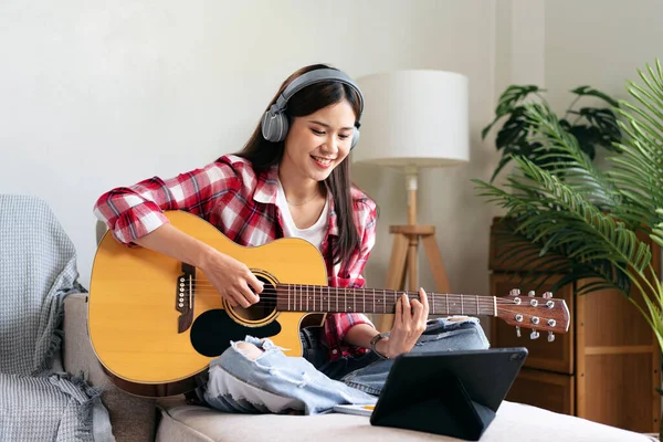 Young woman is playing guitar and practice to singing the song while wearing headphone to watching video tutorial on playing a musical instrument and sitting on comfortable the couch in living room.