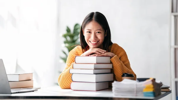 Distance education learning concept, Young woman wearing sweater and smiling with pile of a books while reading a book and studying education lesson online at home.