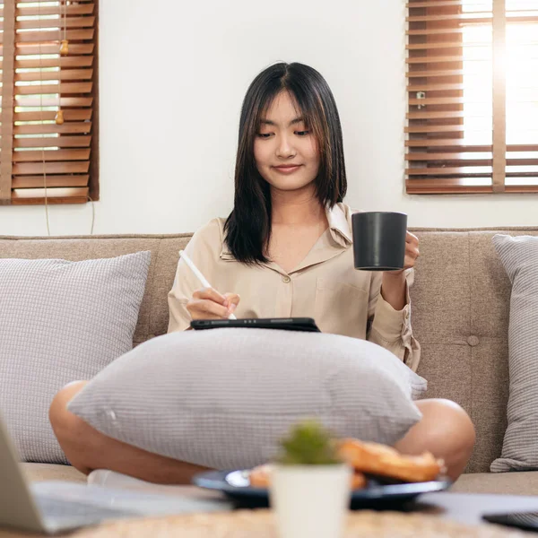 女企业家坐在舒适的大沙发上 把平板电脑放在枕头上 一边写数据 一边喝咖啡 一边在客厅里工作 — 图库照片