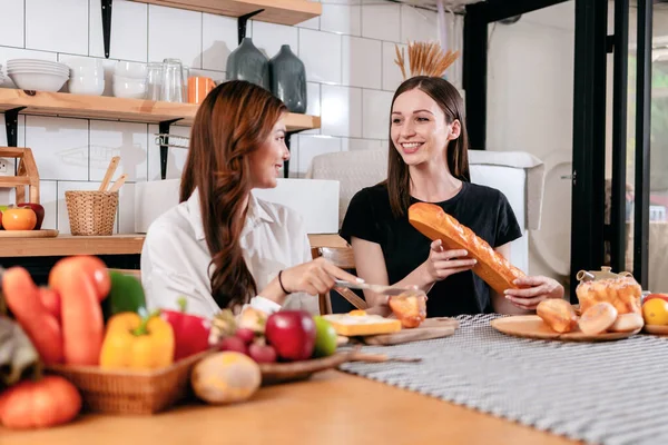 两名年轻女子在木制切菜板上切面包 并在桌上准备新鲜蔬菜和水果 然后在家里的现代厨房为健康的早餐做饭 — 图库照片