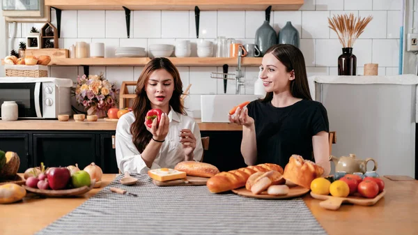 两名年轻女子手里拿着胡椒粉和胡萝卜 一边享用着 一边准备桌上的新鲜蔬菜和水果 一边在家里的现代化厨房里为健康的饭菜做饭 — 图库照片