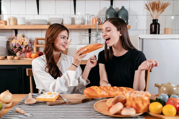 年轻女子一边吃着面包 一边给朋友喂着面包 准备在家里的现代厨房里一起烹调健康的早餐 — 图库照片