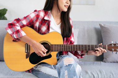 Genç bir kadın, evdeki kanepede rahat bir şekilde otururken gitar ve pop şarkıları çalıyor..