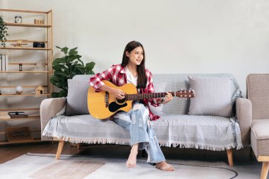 Genç kadın evdeki kanepede rahat bir şekilde otururken gitar çalıyor ve şarkı söylemeye çalışıyor..