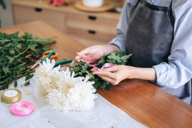 Önlüklü kadın çiçekçi pembe kurdele kullanarak müşteriye teslim etmek için çiçek dükkanında beyaz kasımpatı buketi yapıyor..