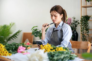 Önlüklü kadın çiçekçi, çiçekçi dükkanında sarı kasımpatı ile çiçek buketi yaparken ciddi bir yüzle akıllı telefonda düzenin detaylarını okuyor..