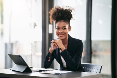 Takım elbiseli Afrikalı Amerikalı iş kadını dijital tablet kullanarak evrak işleriyle ilgili muhasebe bilgilerini okurken modern çalışma alanındaki yeni iş projelerinin planlama ve stratejisini düşünüyor..