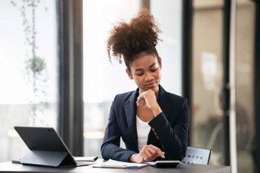 Takım elbiseli Afrikalı Amerikalı iş kadını, iş hesaplarını hesaplamak ve modern çalışma ortamında dijital tablet ve evrak işleriyle uğraşırken proje planlamasını hesaplamak için hesap makinesi kullanıyor..