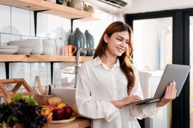 Ev eğlencesi yaşam tarzı konsepti. Mutfak odasında sıradan giyinmiş, dizüstü bilgisayarı olan genç bir kadın. Evde boş zamanlarında arkadaşlarıyla yazışıp sohbet ediyor..