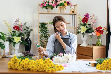 Önlüklü kadın çiçekçi konuşmak için akıllı telefon kullanırken müşteriyle siparişi alırken aynı zamanda kasımpatı sapını keserek çiçekçisinde çiçek buketi yapıyor..