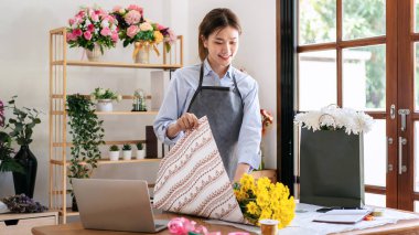 Çiçekçi dükkanındaki çiçek buketini düzenlemek ve tasarlamak için sarı kasımpatıyı masaya sarmak için el işi kağıt kullanan önlüklü bir kadın..