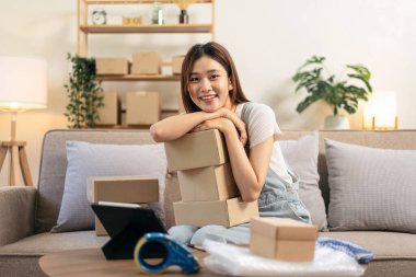 Genç girişimci elinde karton kutular tutarken ve müşteri verilerini tabletten okuyarak müşterilere ev teslimatı için adres yazarken ana ofisteki koltukta rahat bir şekilde oturuyordu..