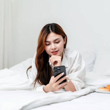 Ev eğlencesi yaşam tarzı konsepti, akıllı telefon kullanarak sosyal medyada gezinen ve arkadaşlarıyla sohbet eden rahat bir yatakta rahat bir yaşam tarzı için evde uzanan günlük kıyafetler giyen genç bir kadın..