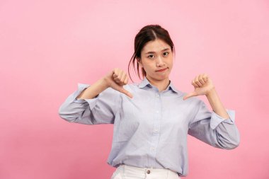Asyalı genç bir kadın ellerini ve parmaklarını kullanarak pembe arka planda hayal kırıklığına uğramış yüzünü göstererek hoşlanmama ya da hoşlanmama sembolünü işaret ediyor..