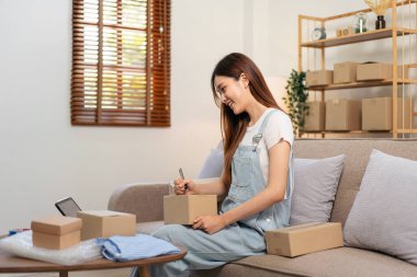 Genç girişimci online siparişleri kontrol etmek için tablet kullanıyor ve karton kutulara müşteri adresleri yazarken aynı zamanda kanepede oturuyor ve ev ofisinin oturma odasında çalışıyor..