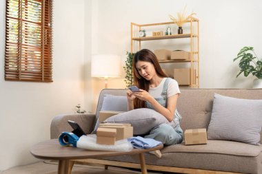 Genç girişimci akıllı telefondan online siparişleri kontrol ediyor ve karton kutulara müşteri adreslerini yazarken aynı zamanda kanepede oturuyor ve ev ofisinin oturma odasında çalışıyor..