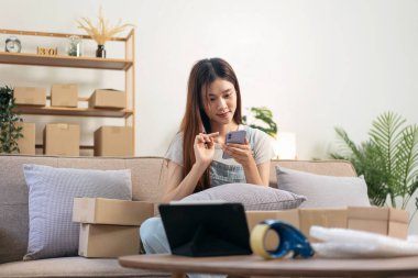 Genç girişimci akıllı telefondan online siparişleri kontrol ediyor ve karton kutulara müşteri adreslerini yazarken aynı zamanda kanepede oturuyor ve ev ofisinin oturma odasında çalışıyor..