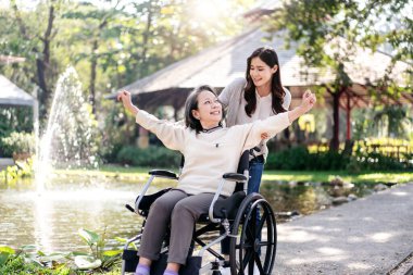 Tekerlekli sandalyedeki son sınıf öğrencisi ve kız aile kavramı, genç Asyalı kadın annesinin tekerlekli sandalyesini bahçede rahatlatırken, son sınıf öğrencisi kadın da kollarını kaldırıp temiz havanın tadını çıkarıyor ve konuşuyor..