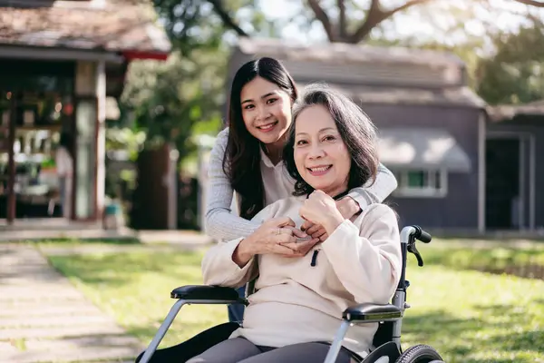 車椅子と娘の家族コンセプトのシニア 若いアジアの女性は 庭で一緒にシニア女性と娘のリラクゼーションしながら 車椅子に障害のある母親を微笑み 抱きしめています — ストック写真