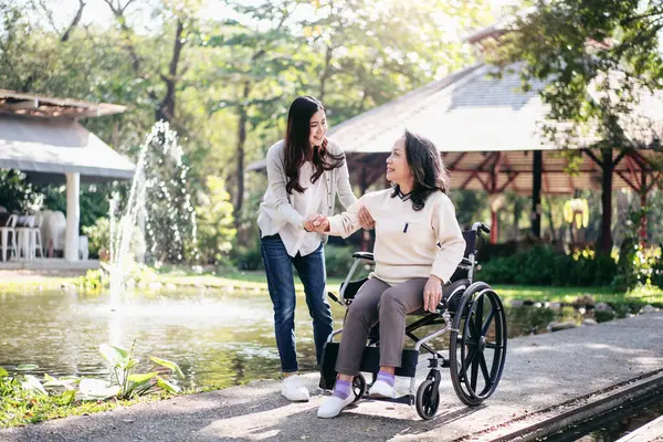 Senior on wheelchair and daughter family concept, Young asian woman helping disabled mother to get up from wheelchair while senior woman and daughter relaxation with enjoying fresh air in the garden.
