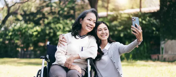 Senior on wheelchair and daughter family concept, Young asian woman embracing and selfie on smartphone with disabled mother on wheelchair while senior woman and daughter relaxation together in garden.